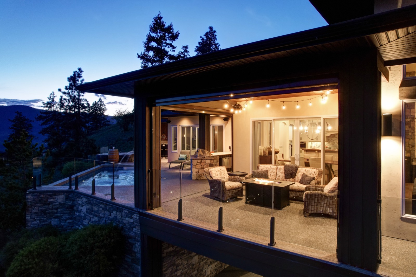 A covered outdoor living area at dusk with string lights, a fire table, wicker seating and a pool beyond the glass railing.