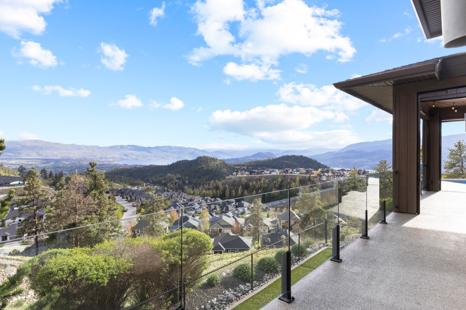 A glass-railed deck looking out over a hillside neighbourhood toward the lake and mountains on a clear day.