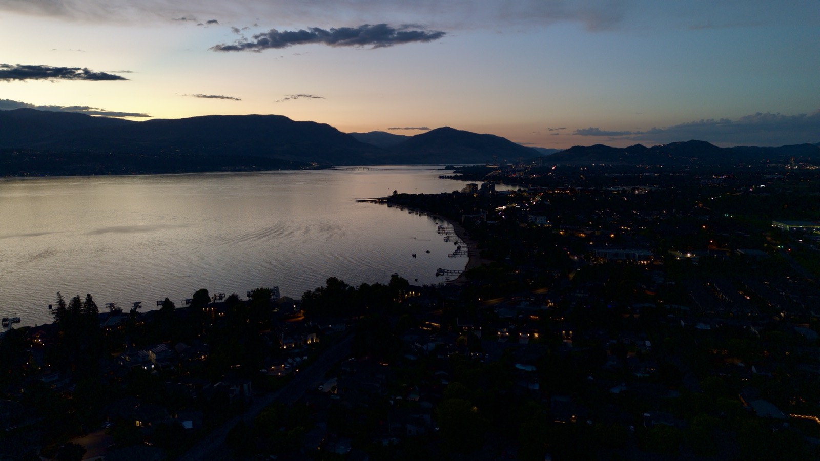 An aerial view of the Kelowna waterfront after sunset, the lake reflecting the last light with city lights coming on along the shore.