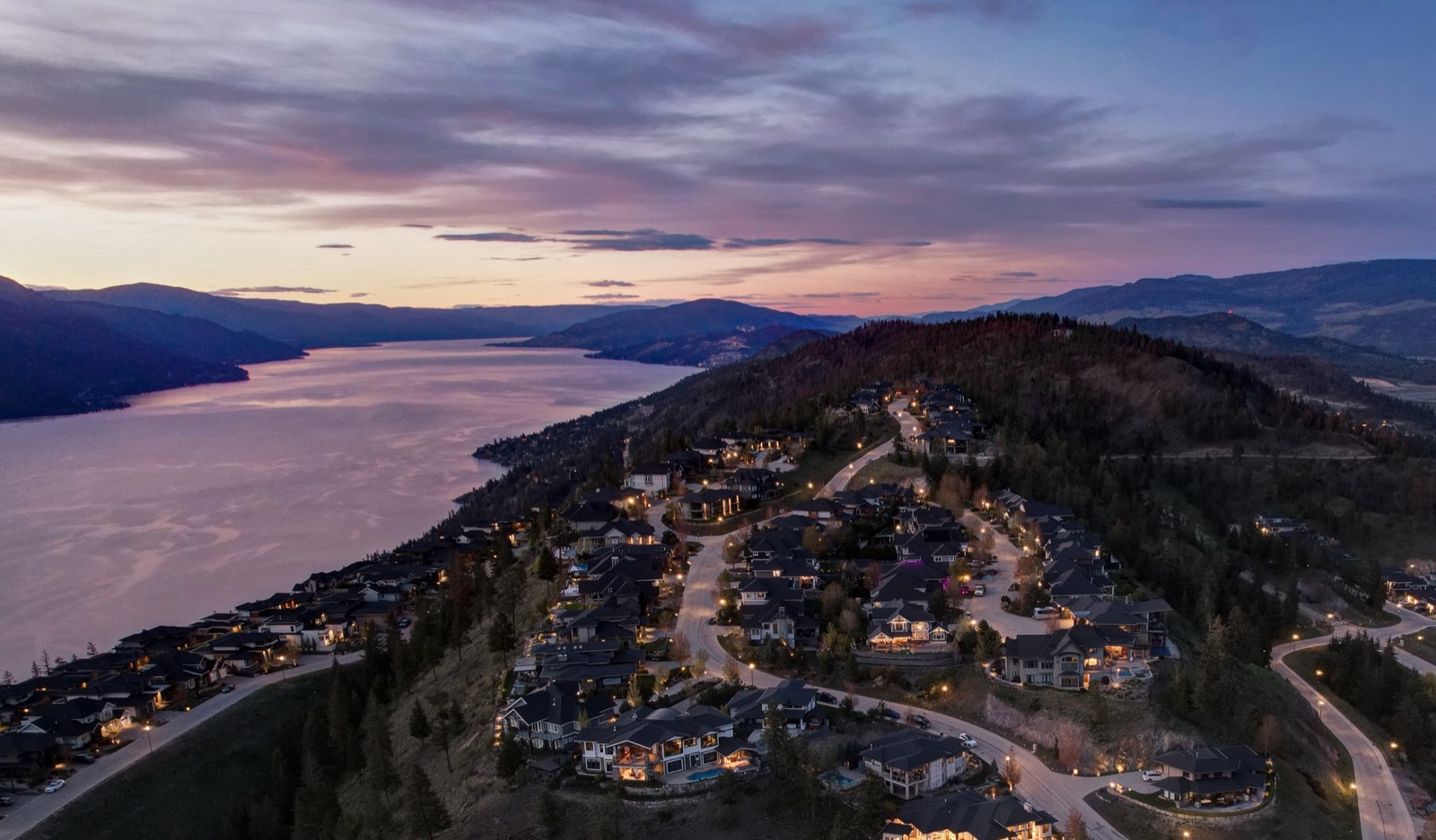 An aerial view at dusk of a hillside neighbourhood above Okanagan Lake, streetlights and windows lit along winding roads.