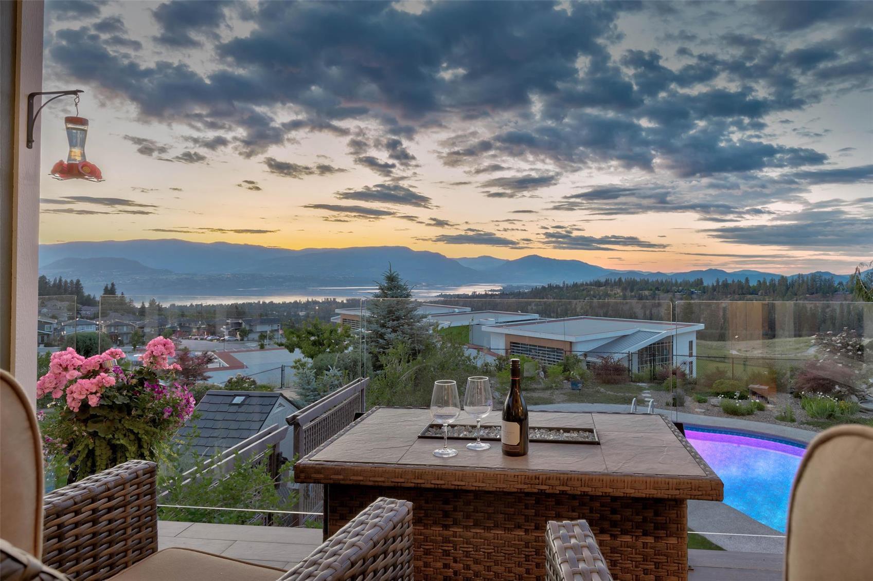 A patio table set with wine glasses at sunset, overlooking a lit pool, the valley and Okanagan Lake beyond.