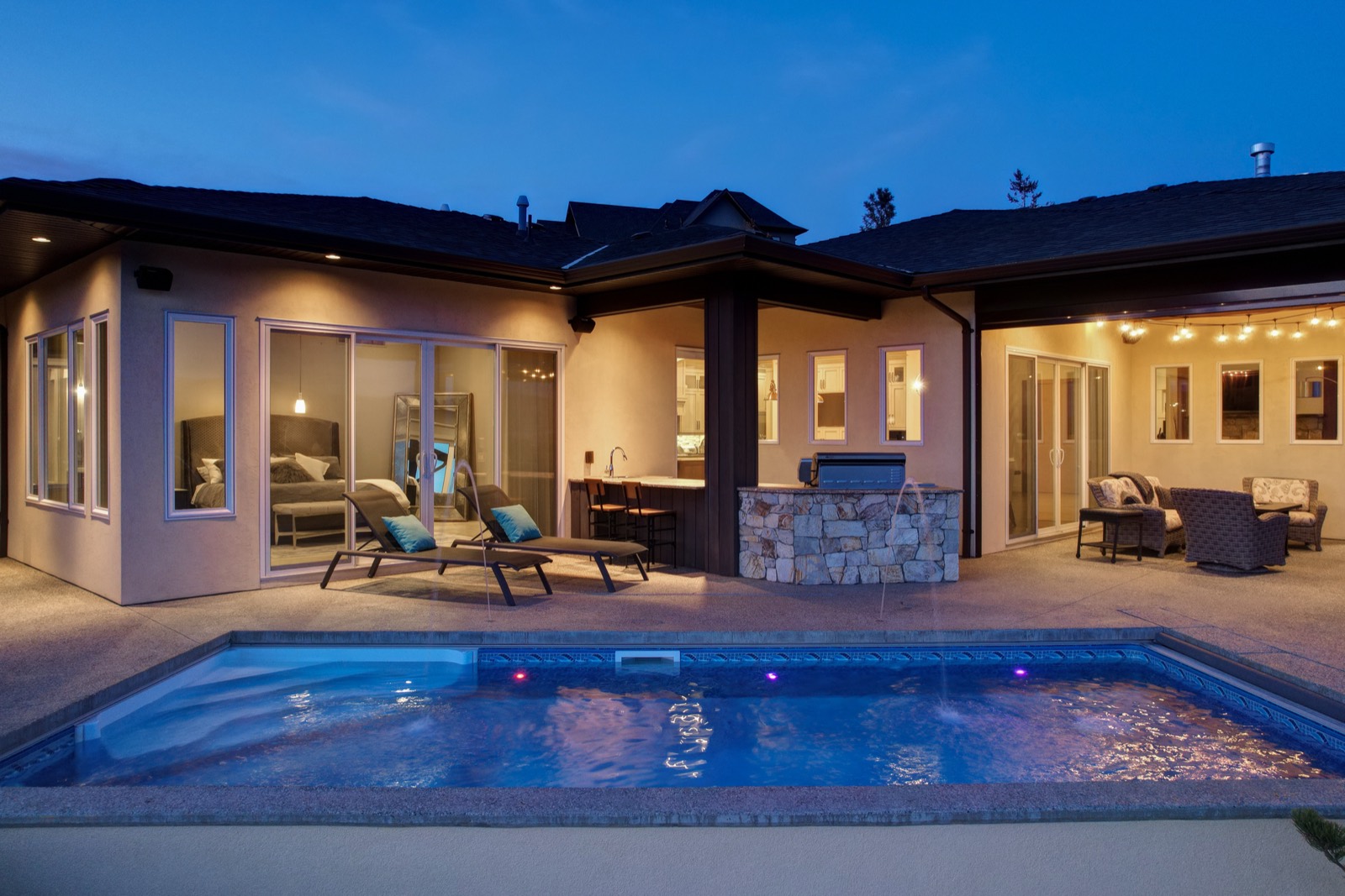 A lit swimming pool and patio behind a single-storey home at blue hour, with the bedroom and outdoor kitchen glowing through open glass doors.