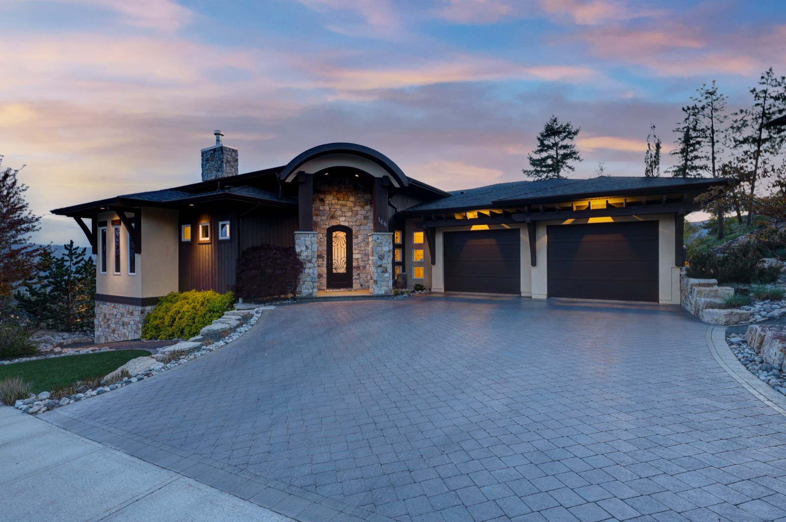 A modern stone-and-timber home photographed at dusk, warm interior lights on and a pink and blue sky overhead.